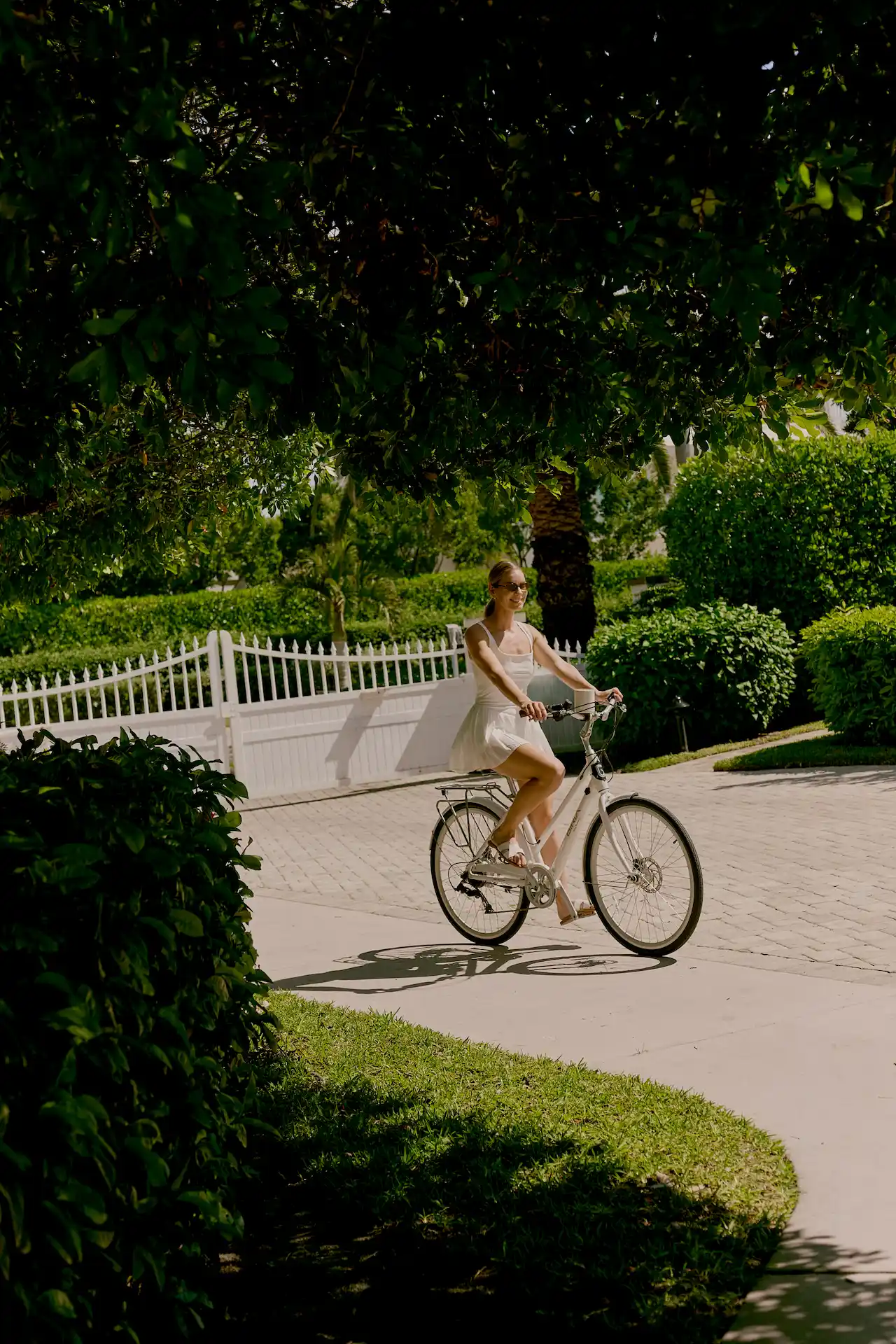 Person riding a white bicycle along a sunlit path surrounded by lush greenery and a white fence
