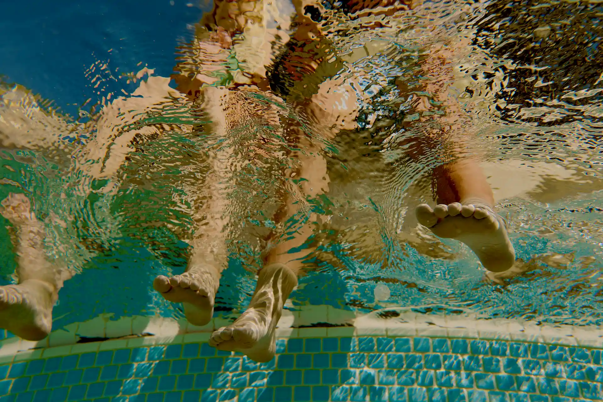 Underwater view of legs and feet dipping into a sunlit pool with playful ripples and mosaic tiles