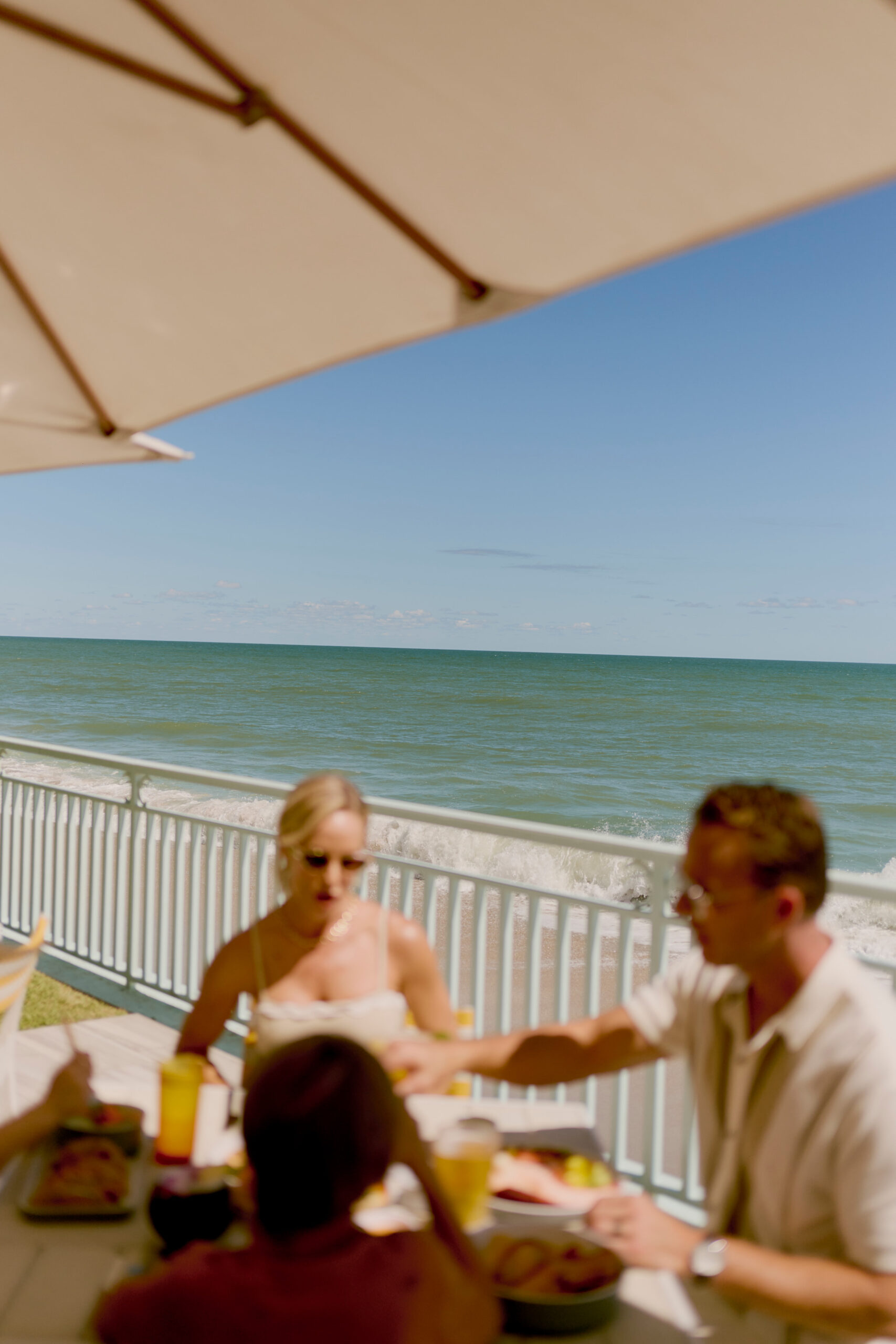 Family dining outdoors under umbrella with ocean view and waves in background