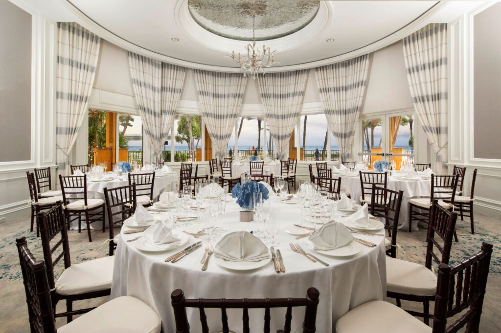 Elegant oceanview banquet room with round tables, blue centerpieces, and tall draped windows