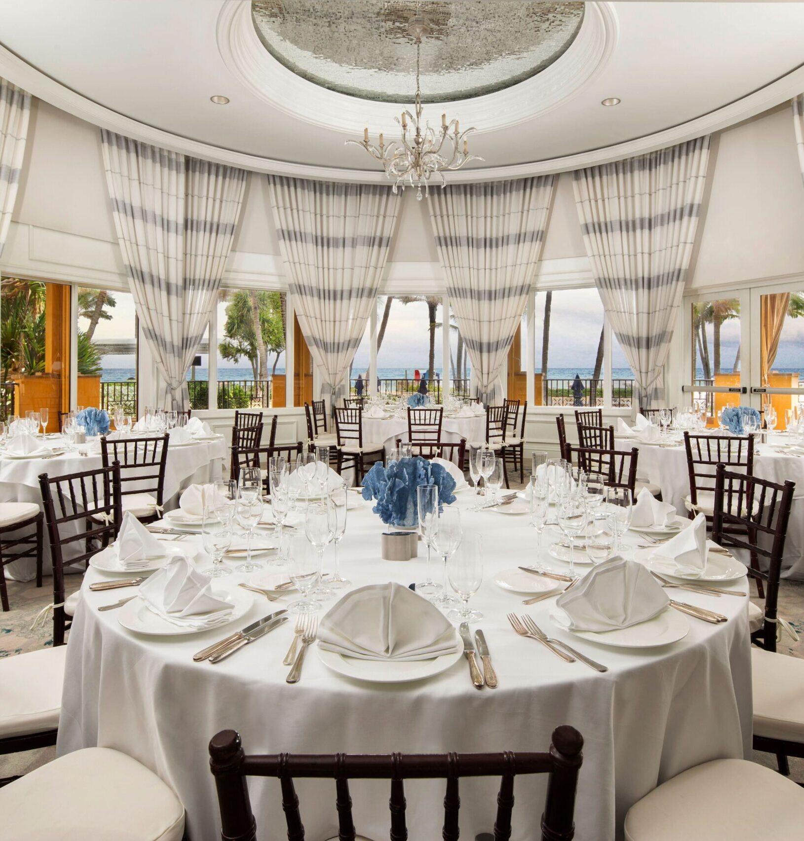 Elegant oceanview banquet room with round tables, blue centerpieces, and tall draped windows
