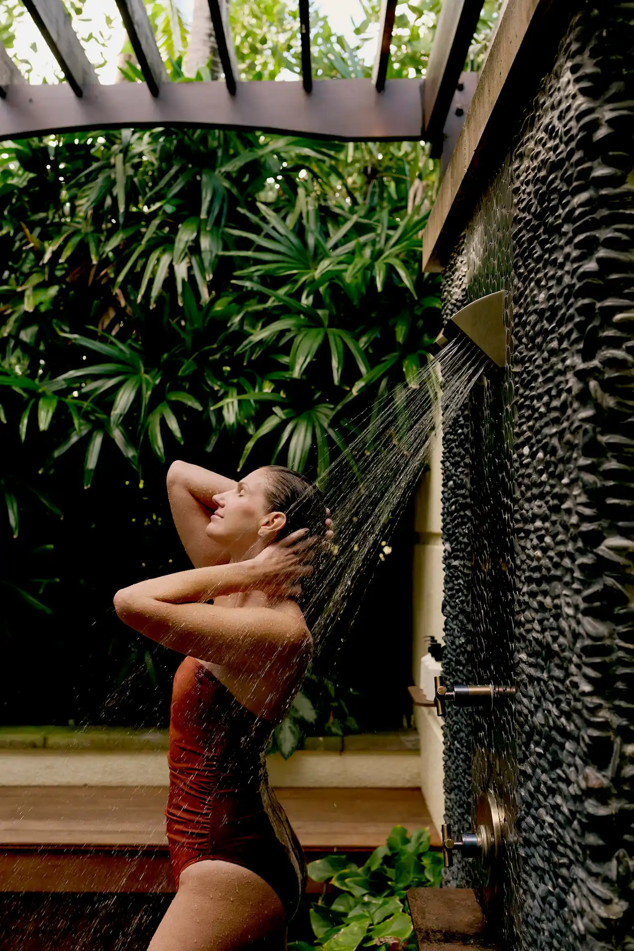 Person rinsing off under an outdoor shower surrounded by lush tropical greenery and warm natural light