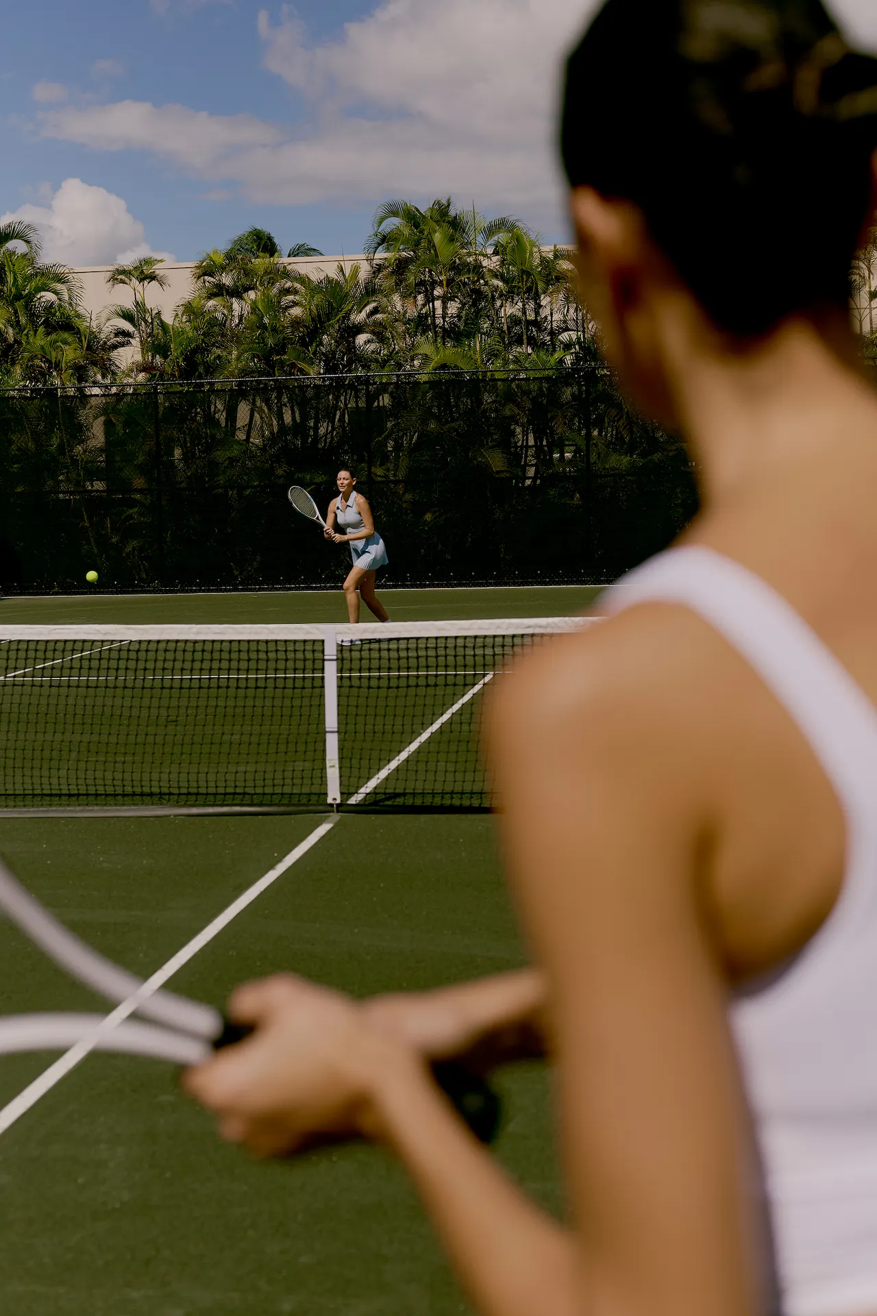 Two people playing tennis on an outdoor court surrounded by palm trees under a partly cloudy sky
