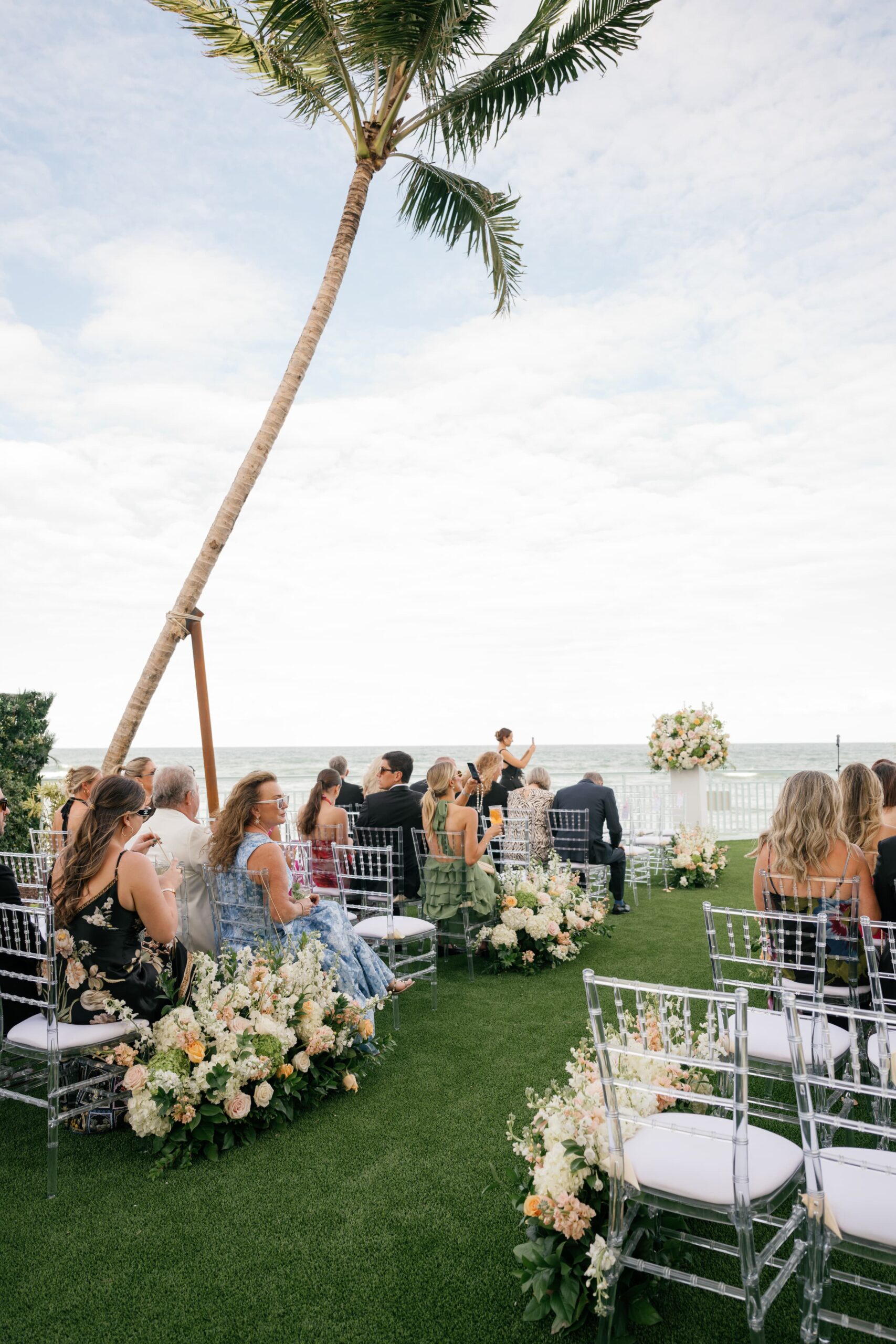 Guests seated at oceanfront wedding ceremony with floral arrangements and palm tree backdrop