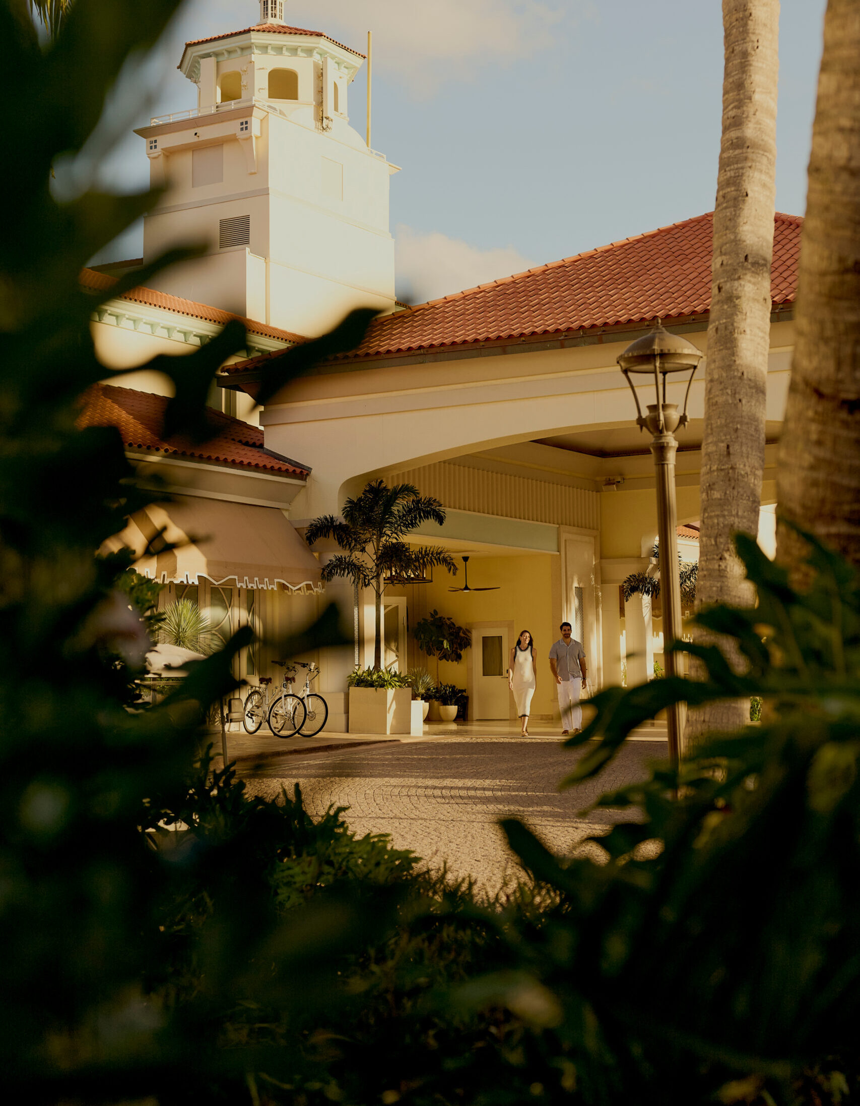 Couple walking toward elegant resort entrance with red tile roof and palm trees at sunset