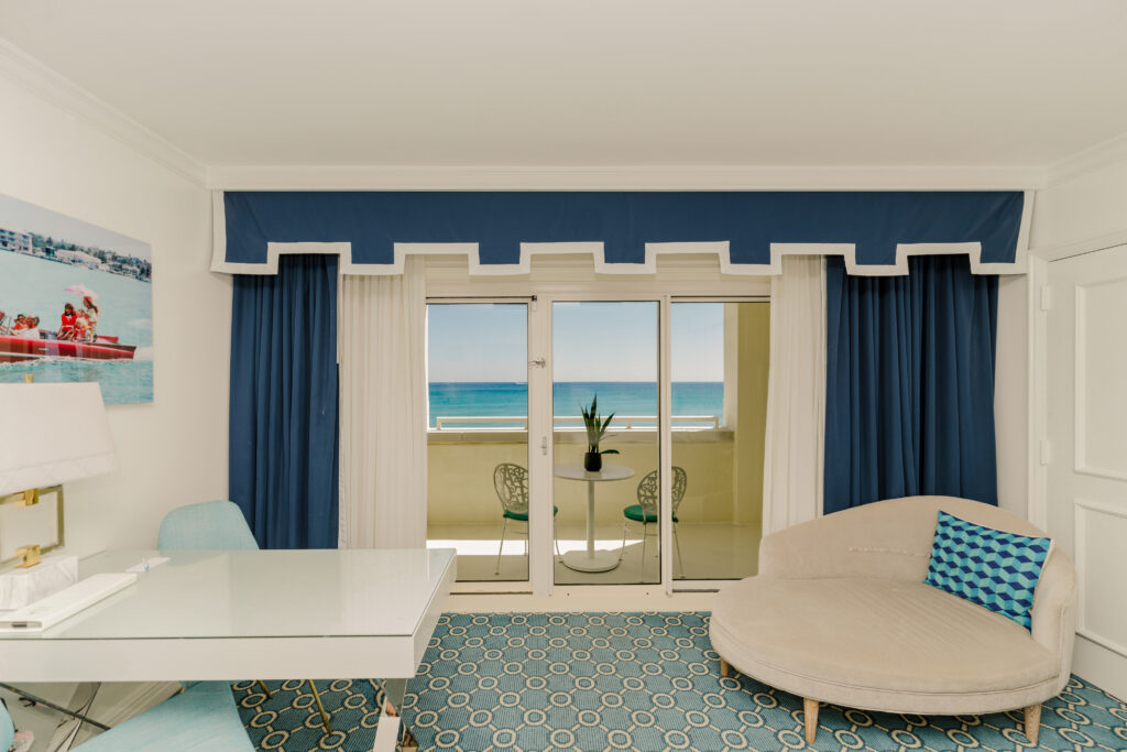 Bright hotel room with blue curtains, desk, sofa, and balcony overlooking turquoise ocean
