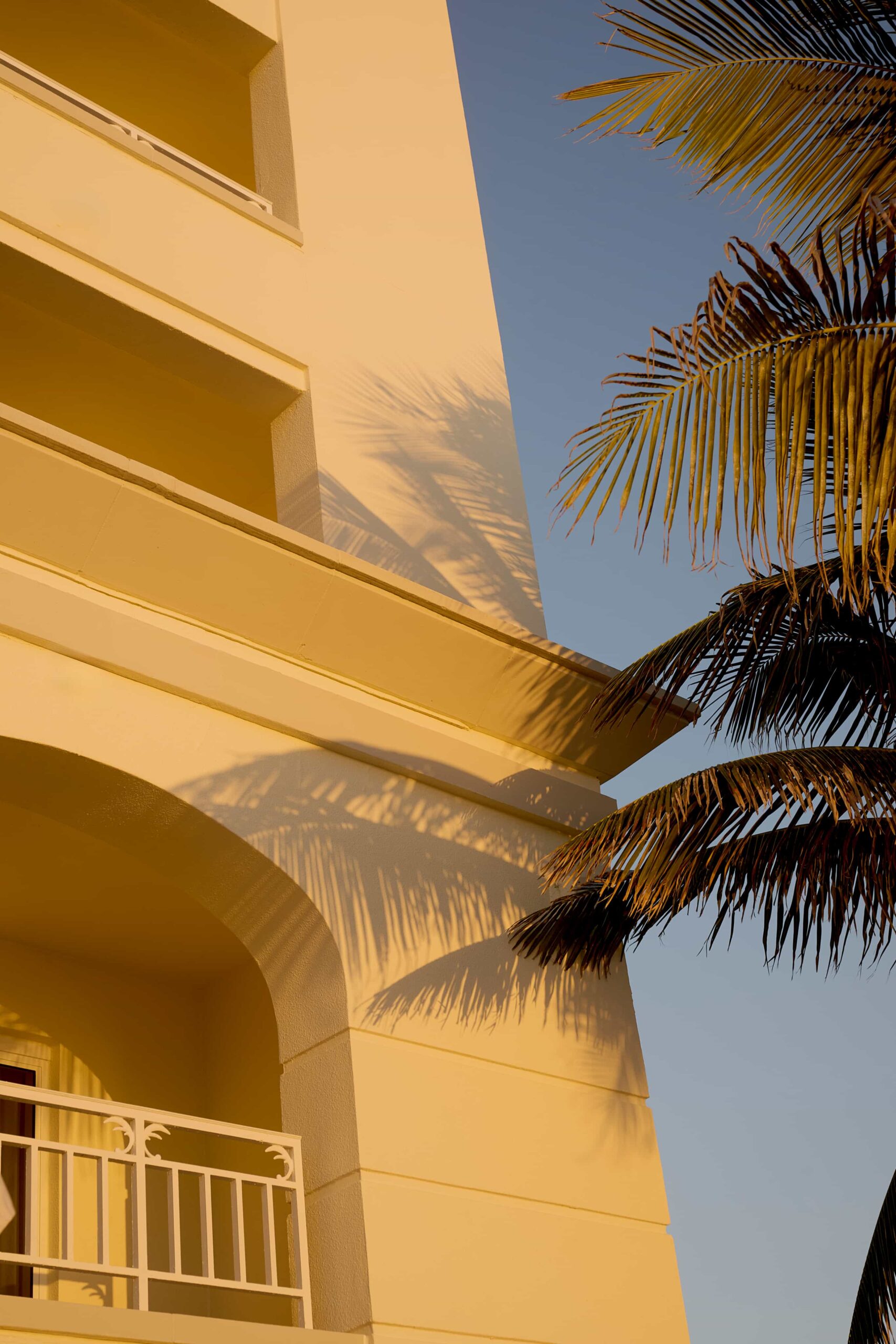Sunlit cream building with balconies and palm tree shadows against clear blue sky