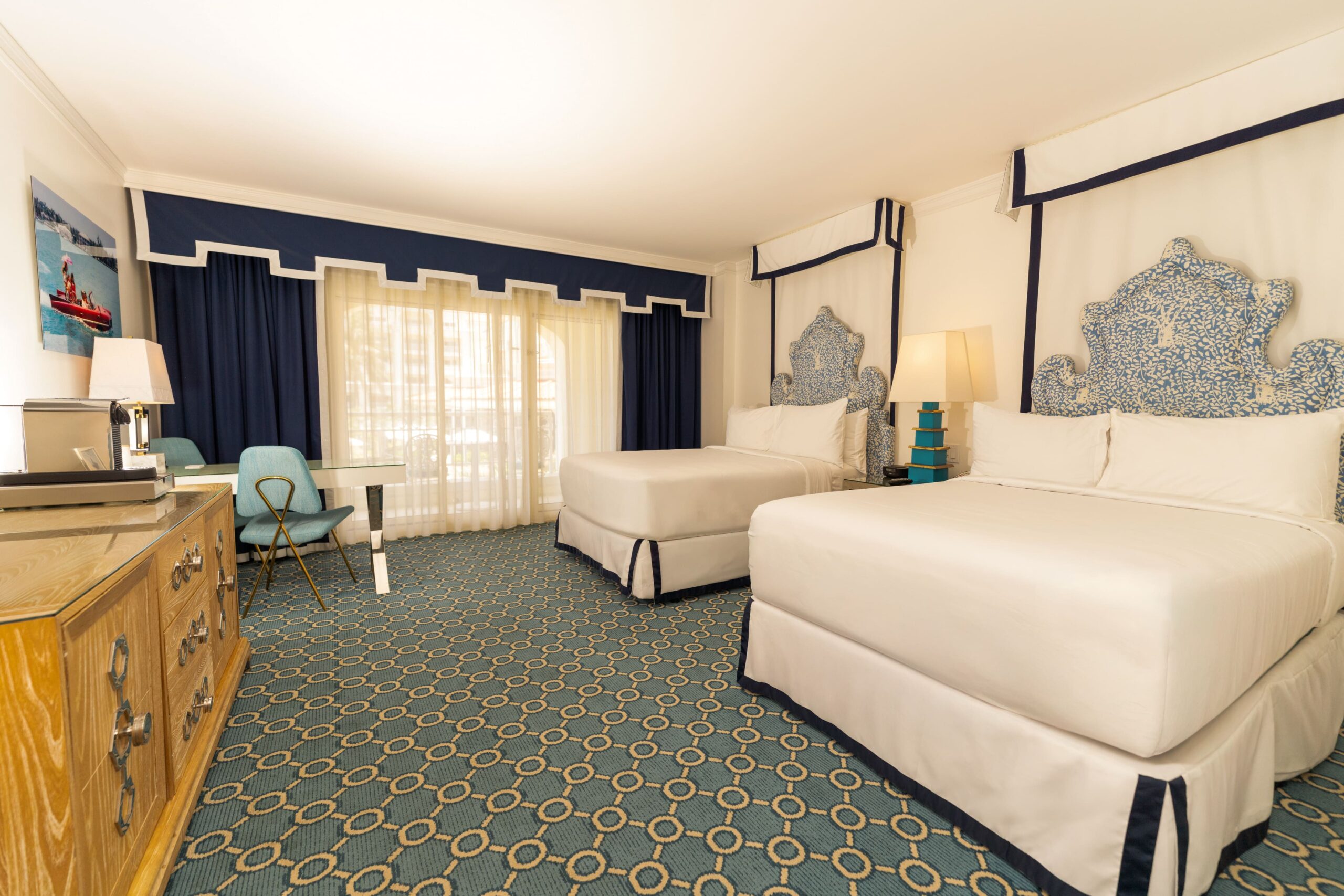 Spacious hotel room with two beds, blue accents, and desk by balcony window