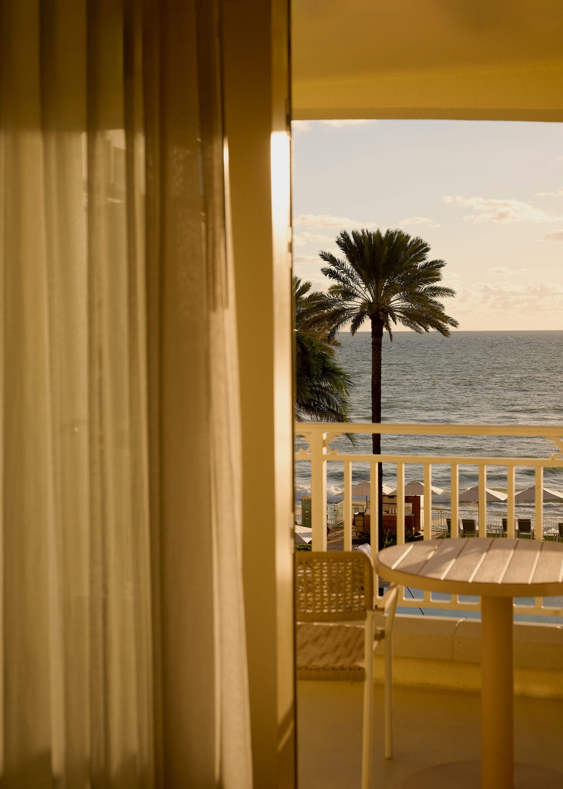 Balcony view of ocean at sunset with palm tree, beach umbrellas, and patio table in warm golden light