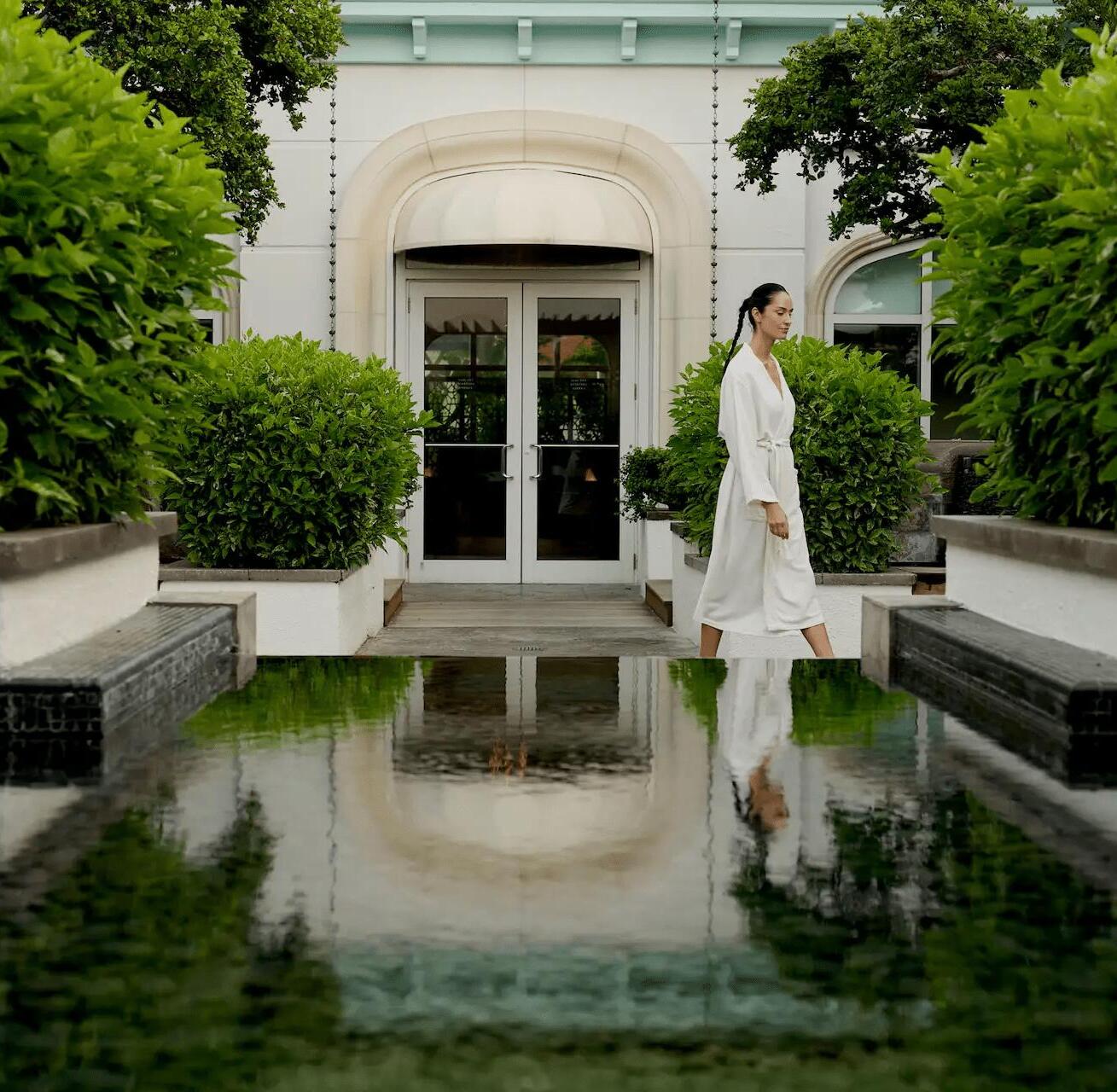 Person in a white robe walking along a tranquil garden path beside a reflective pool at a serene spa setting