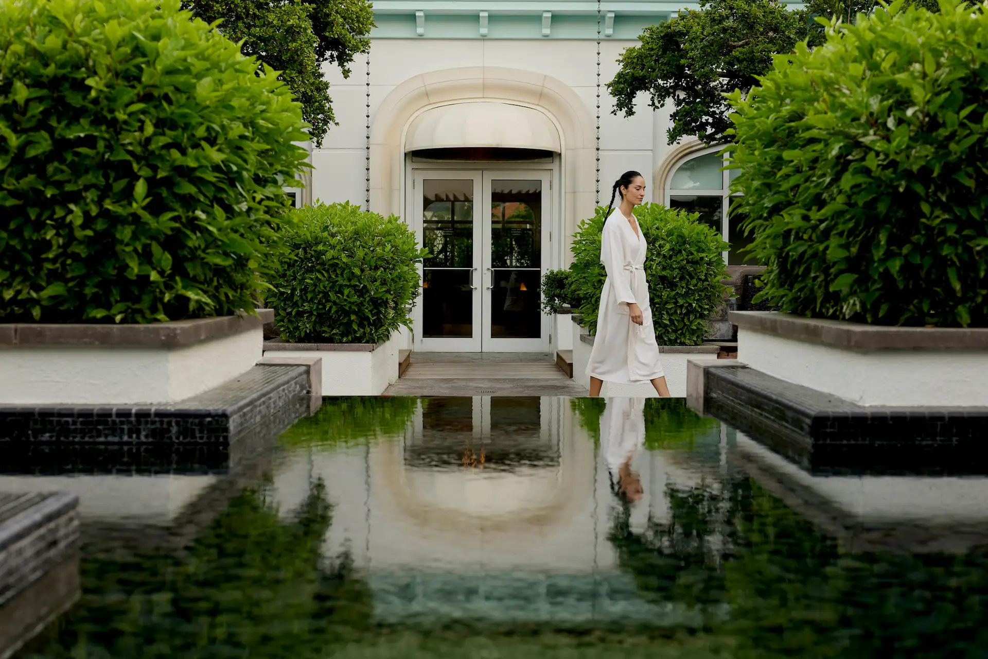 Person in a white robe walking along a tranquil garden path beside a reflective pool at a serene spa setting