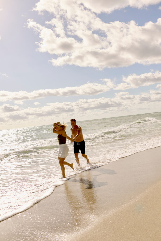 Couple running along shoreline with waves splashing under bright sky