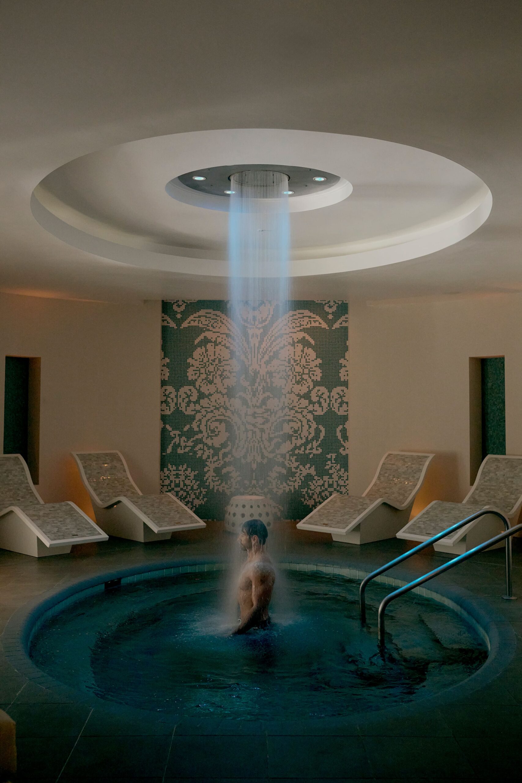 Man standing under waterfall shower in indoor spa pool with lounge chairs