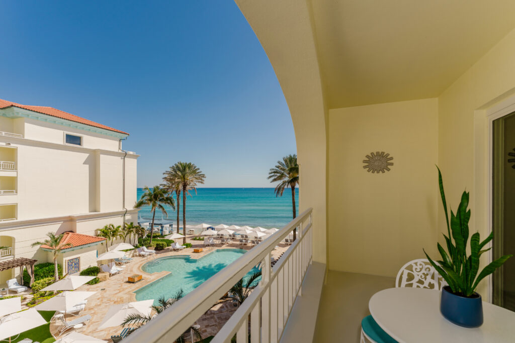 Balcony view overlooking resort pool, palm trees, and turquoise ocean under clear blue sky