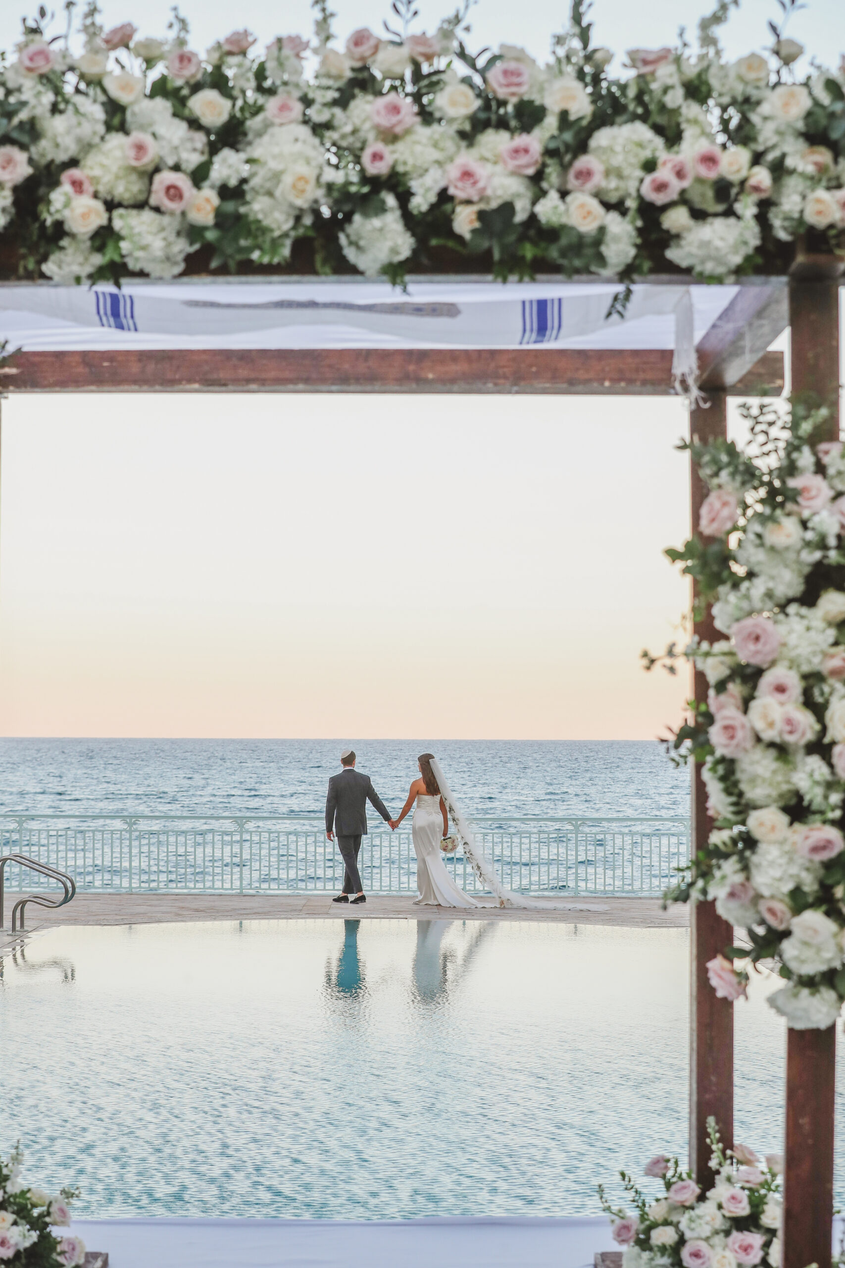 Bride and groom holding hands by oceanfront pool under floral wedding arch