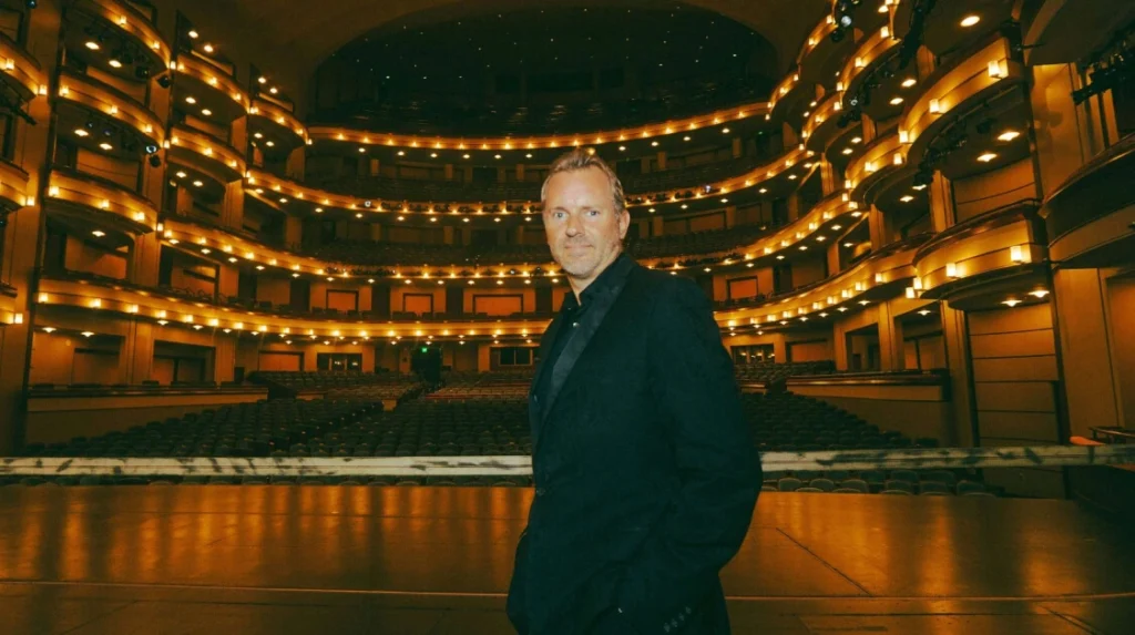 Man standing on a grand theater stage facing empty, ornate balconies under warm lighting.