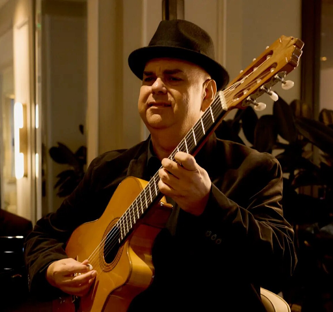 Man in black hat playing acoustic guitar indoors with pianist in background