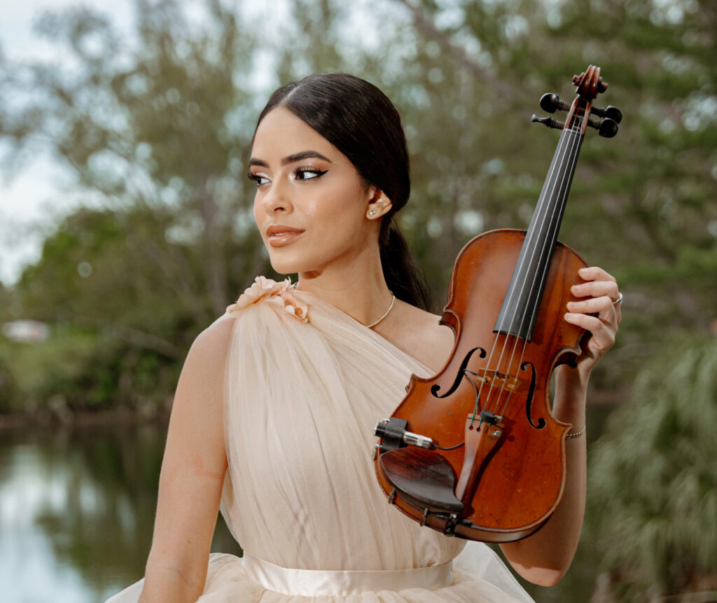 Maggi vi standing on a wooden deck in an elegant cream gown, holding a violin outdoors.