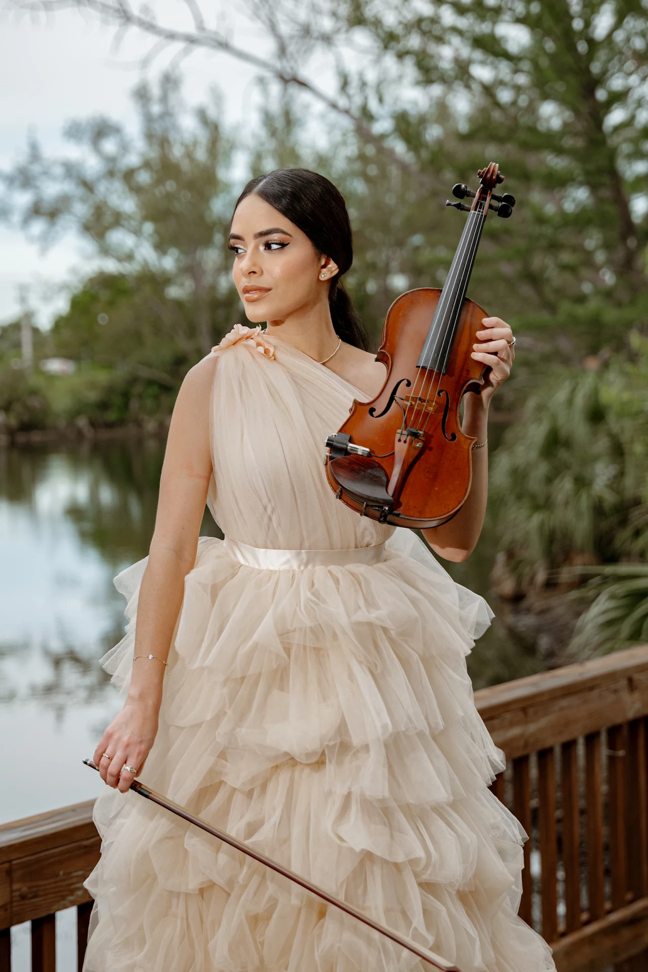 Maggi vi standing on a wooden deck in an elegant cream gown, holding a violin outdoors.