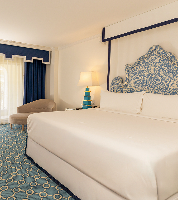 Bright hotel room with white bed, blue patterned headboard, navy curtains, and geometric carpet
