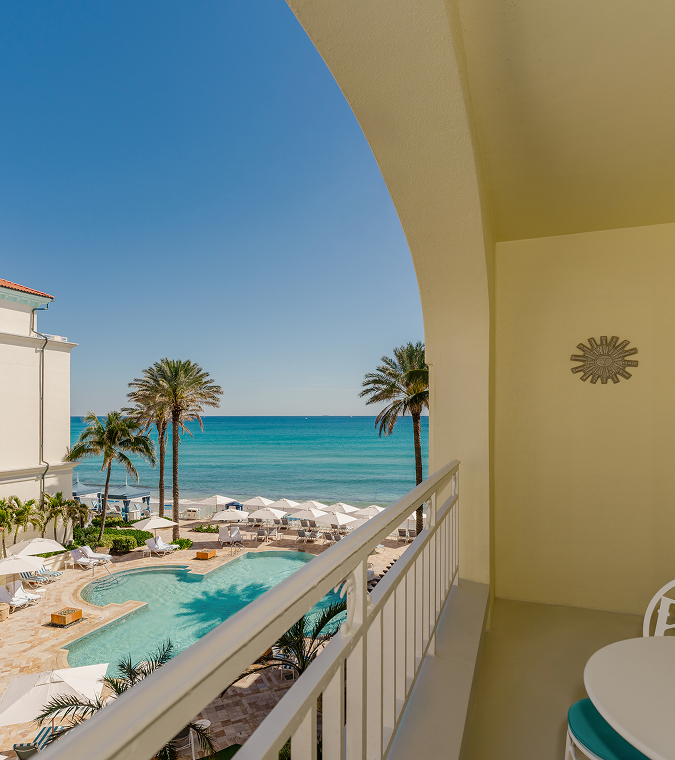 Balcony overlooking turquoise ocean, resort pool, palm trees, and white beach umbrellas