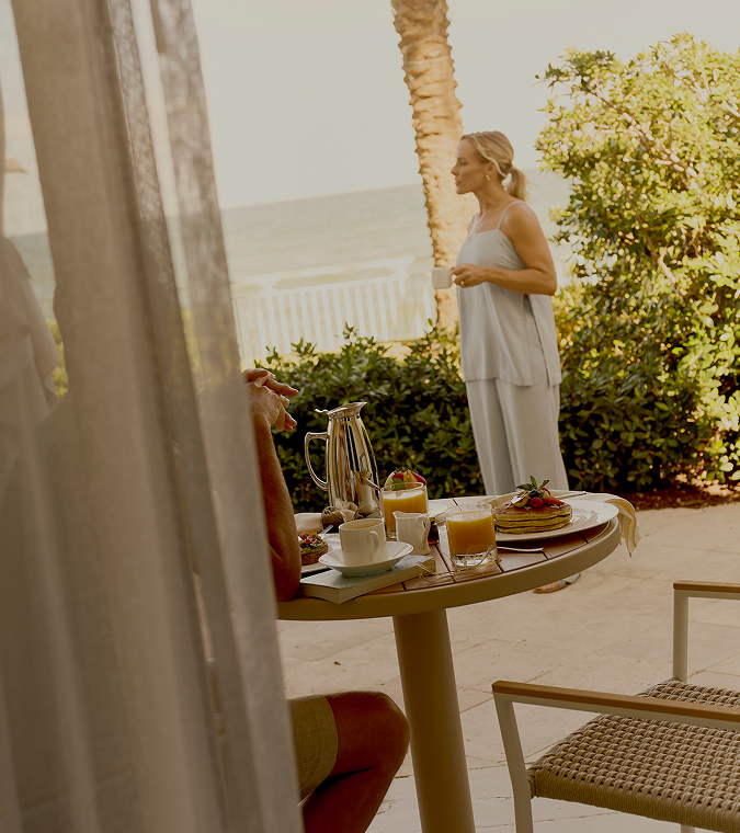 Woman holding coffee on oceanfront patio with breakfast table set with pancakes and juice