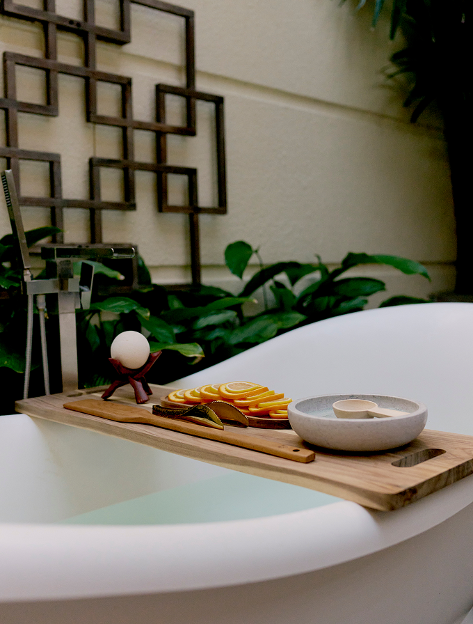 Freestanding bathtub with wooden tray holding citrus slices, bowl, and bath accessory in spa setting