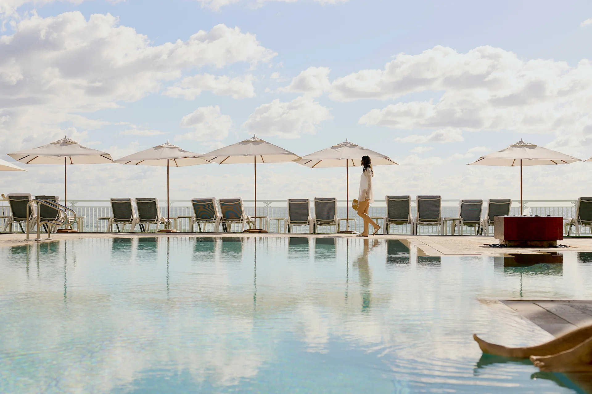 Peaceful poolside scene with white umbrellas, calm water and a guest strolling by the ocean in the background
