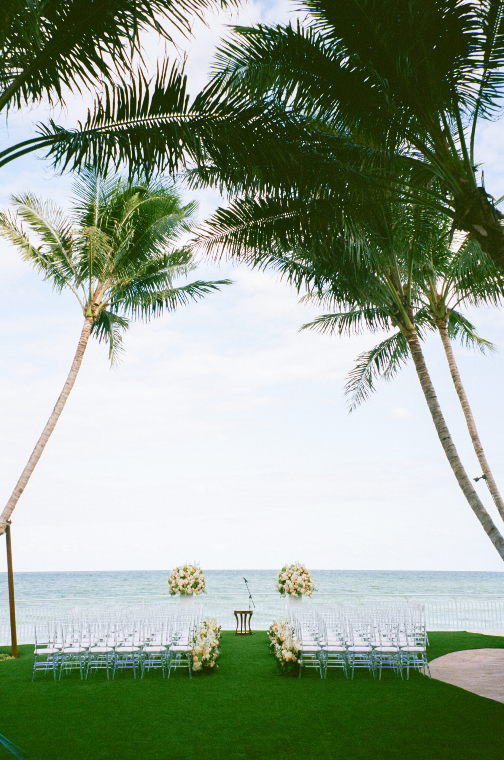 Beachfront wedding setup with white chairs and floral arches beneath palm trees