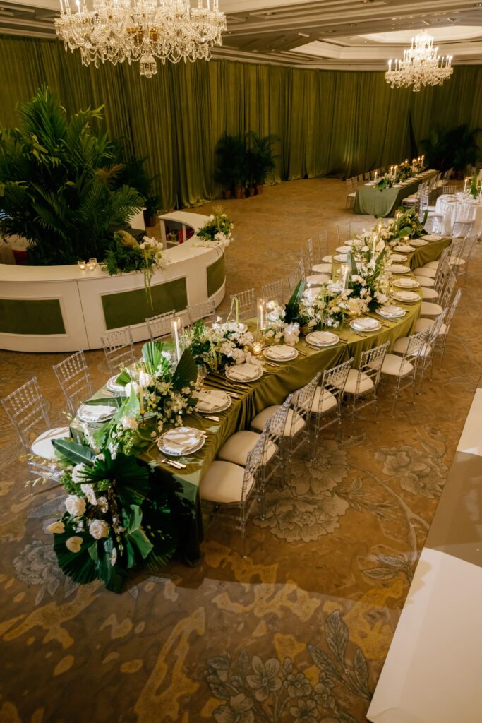 Elegant banquet hall with long floral decorated table, chandeliers, and green draped backdrop