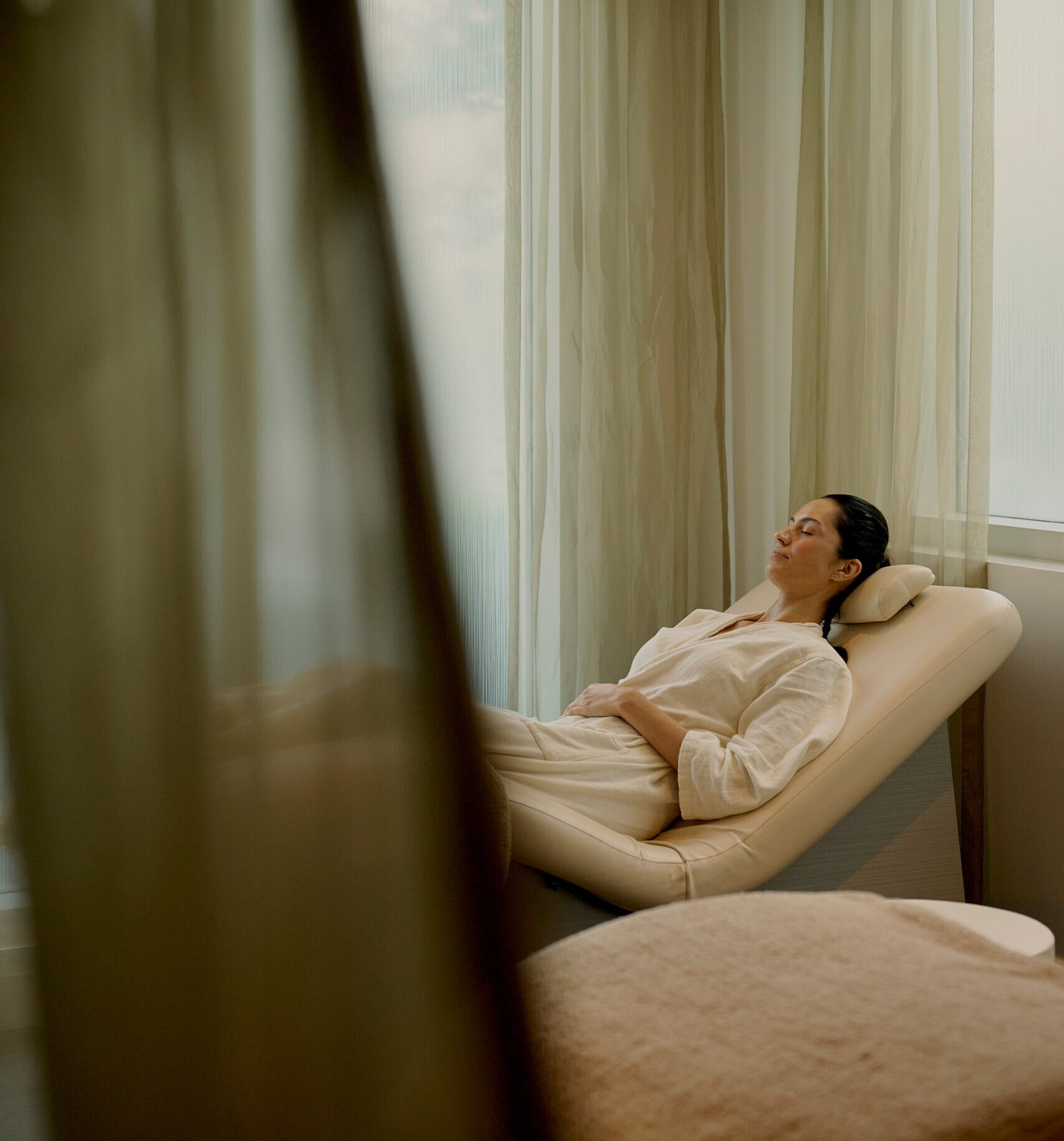 Woman resting on spa lounge chair in softly lit room with sheer curtains