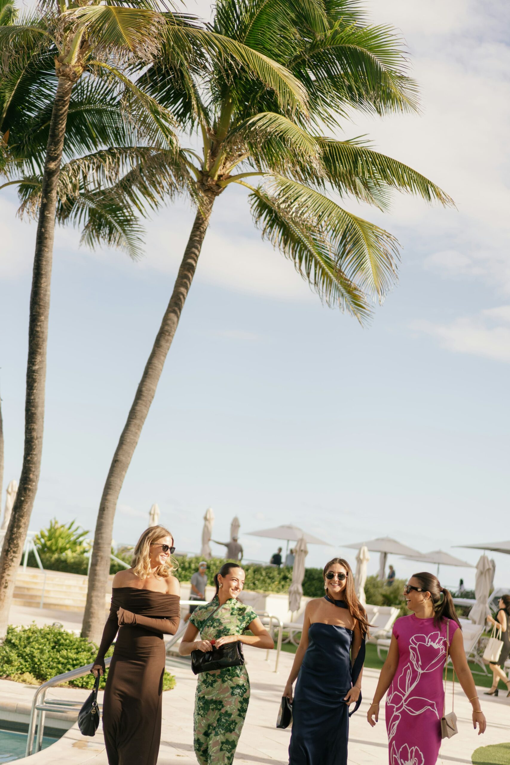 Four women in elegant dresses walking by palm trees at sunny resort pool area
