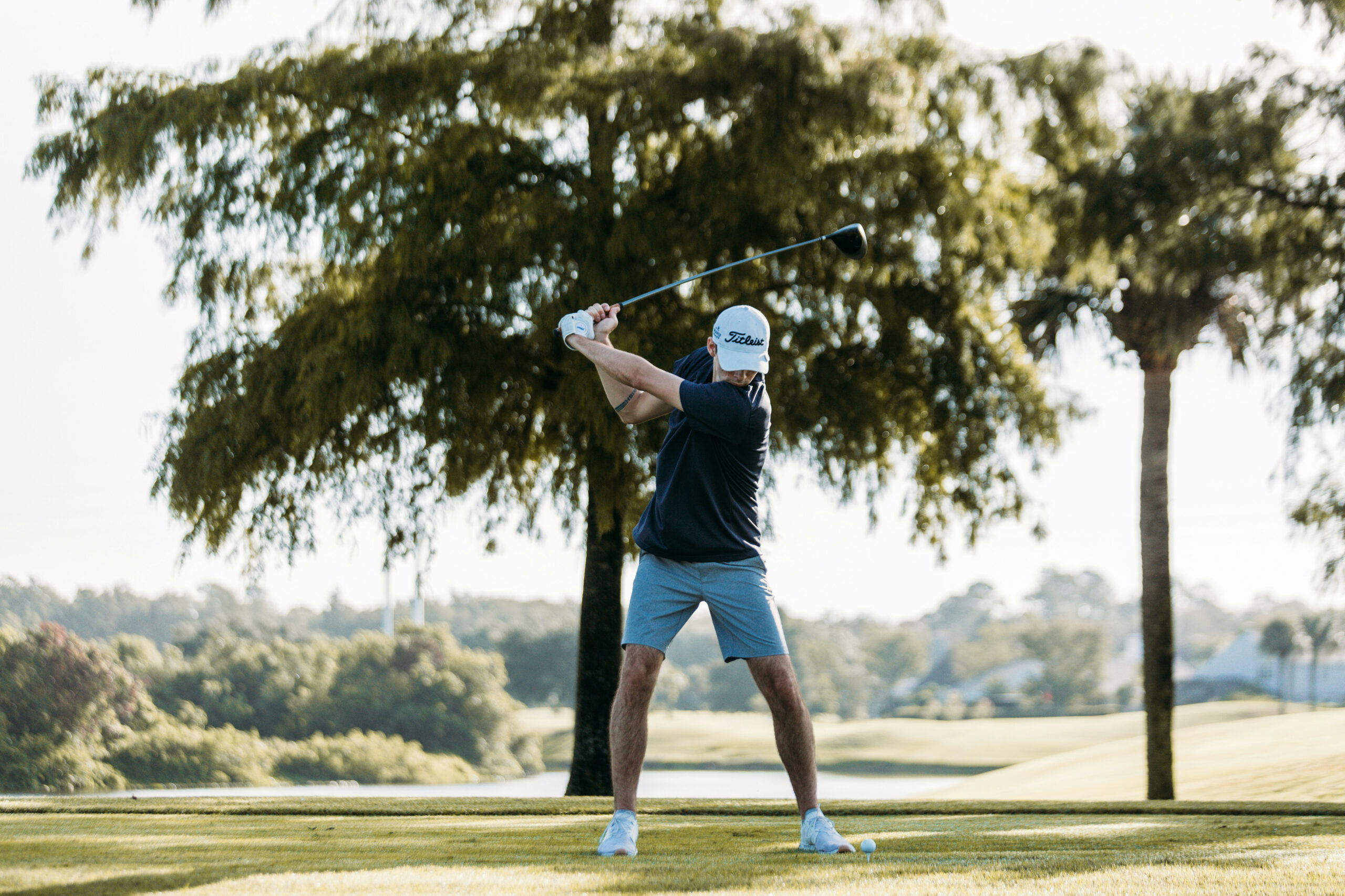 Golfer mid-swing on a sunny course with trees and water in the background