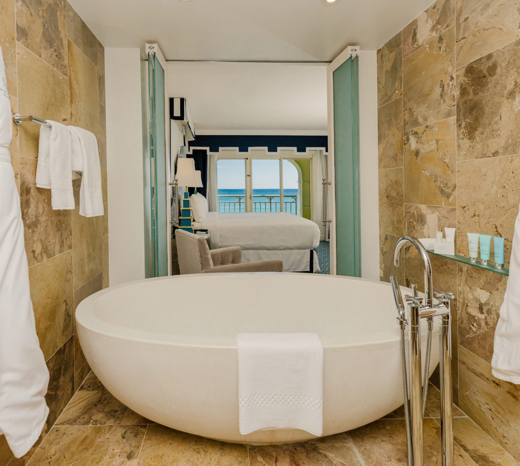 Luxury bathroom with freestanding tub and oceanview bedroom beyond glass doors
