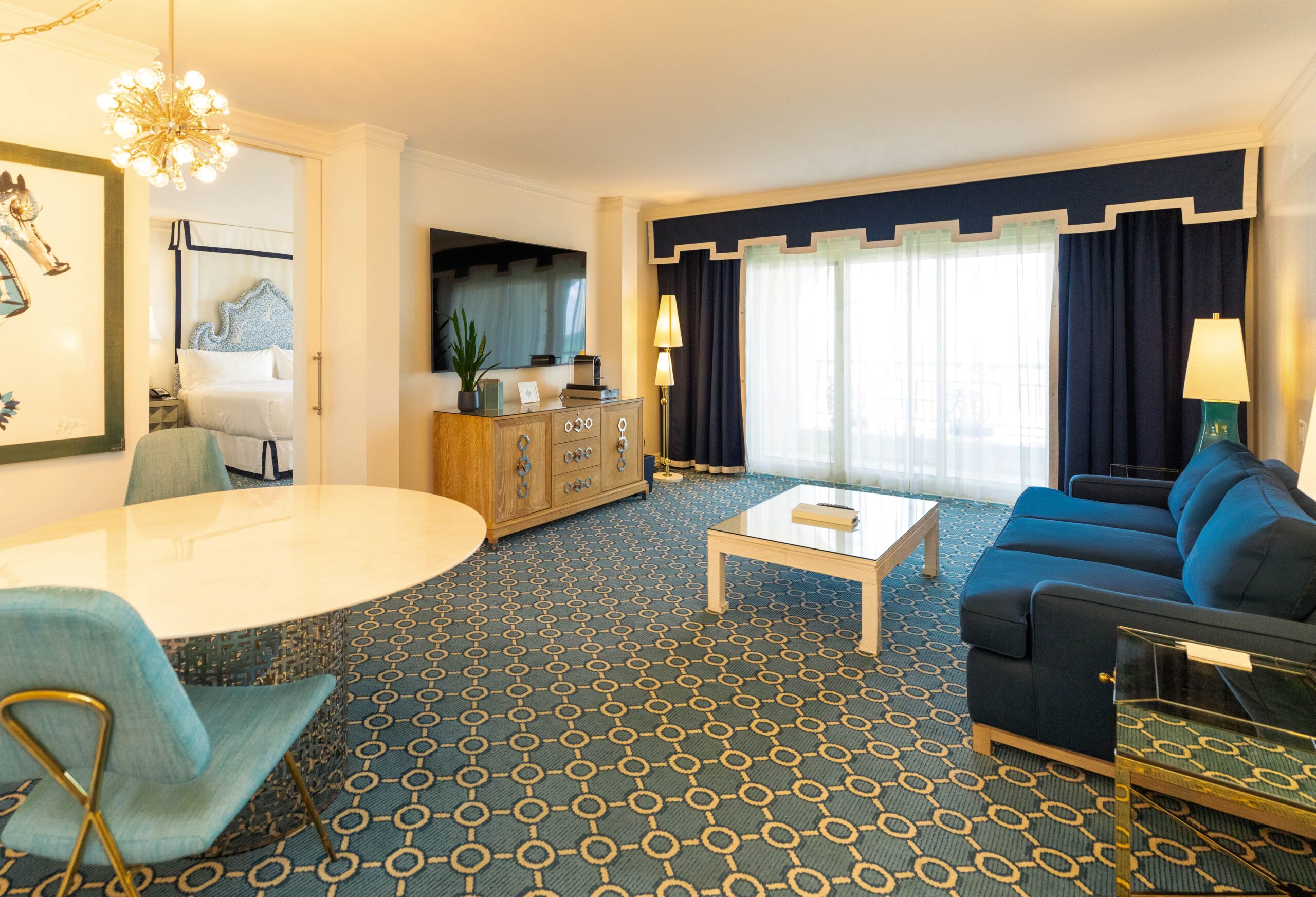 Spacious hotel suite with blue sofa, dining area, and bedroom visible through open doorway