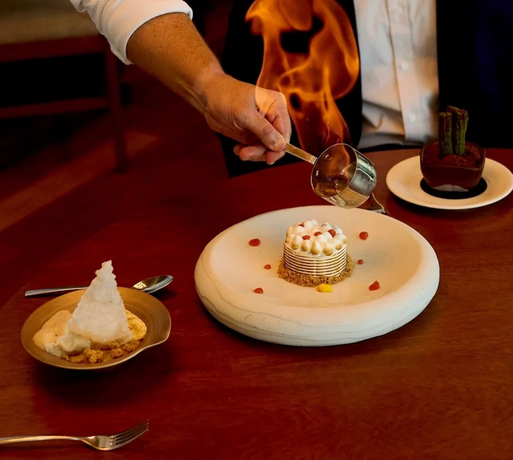 Tableside flambé moment with flames rising over a plated dessert as a guest watches with a smile