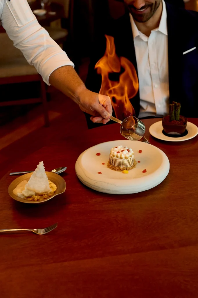 Tableside flambé moment with flames rising over a plated dessert as a guest watches with a smile