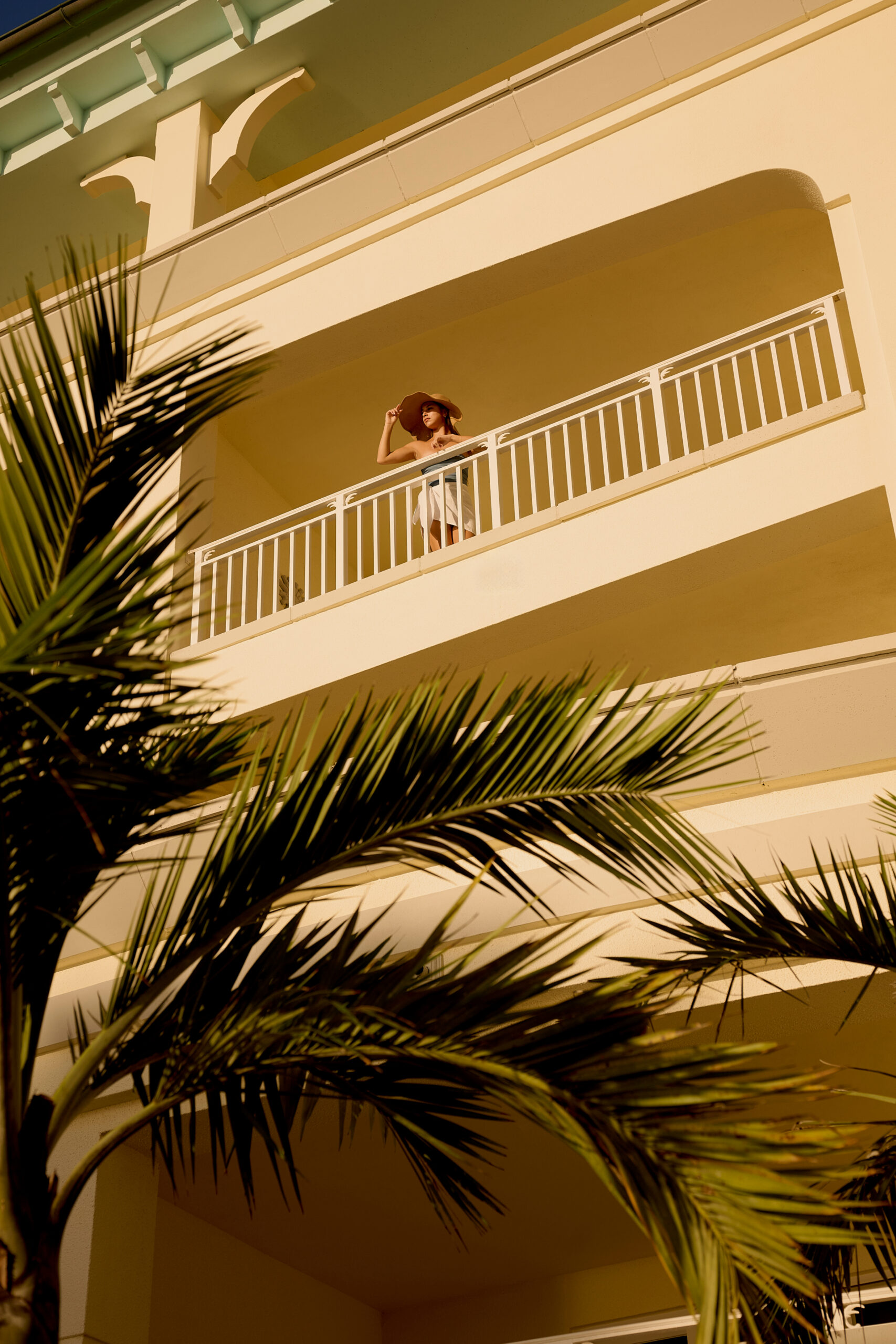 Woman in sun hat standing on resort balcony framed by palm trees