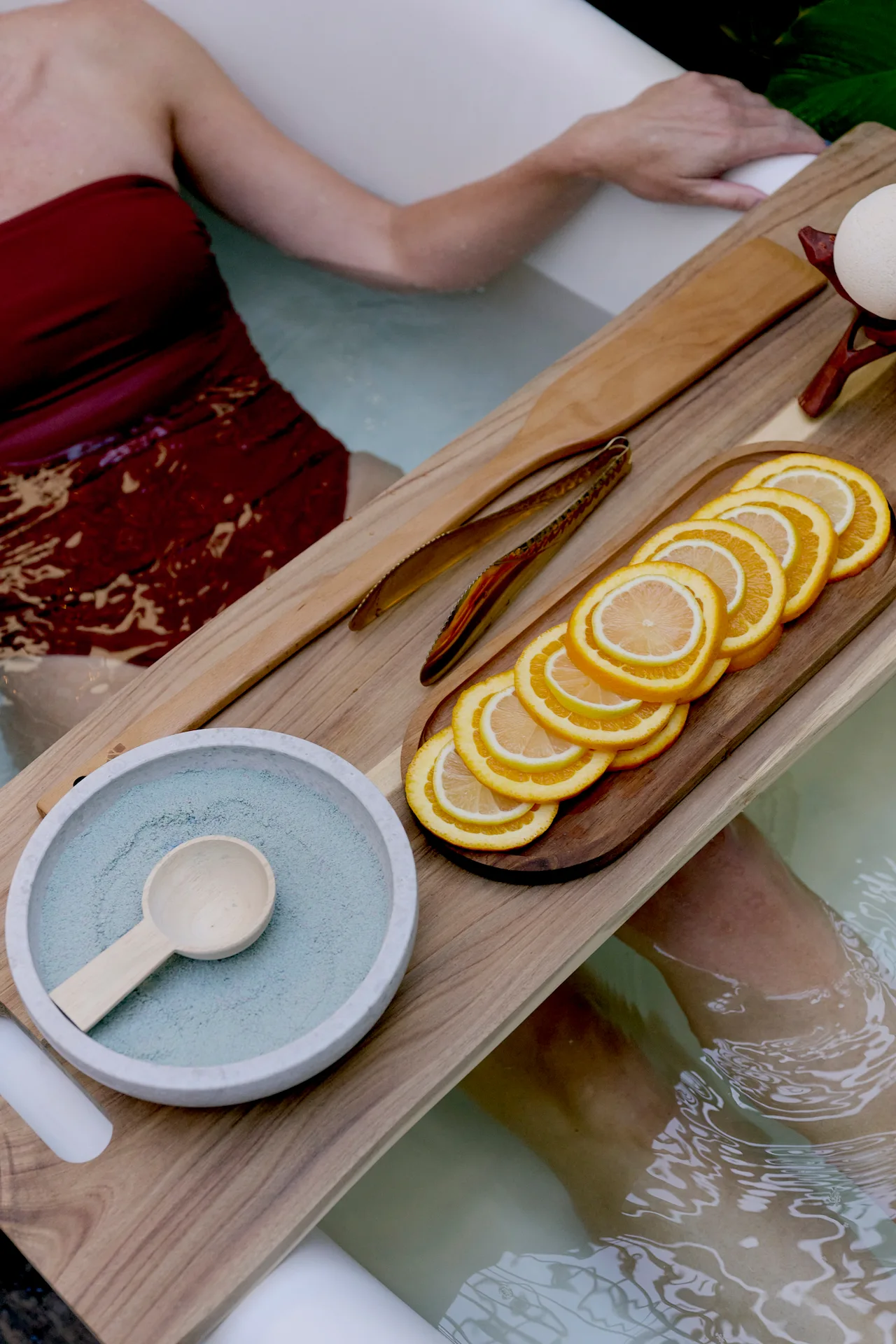 Person relaxing in a warm bath with a wooden tray holding citrus slices, bath salts and spa essentials