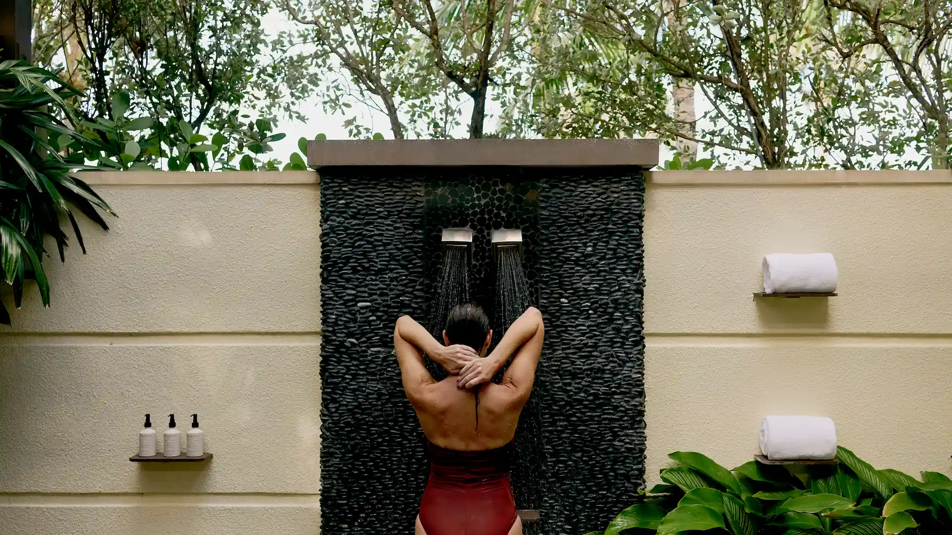 Outdoor shower moment with water streaming down a dark stone wall, surrounded by greenery and soft natural light