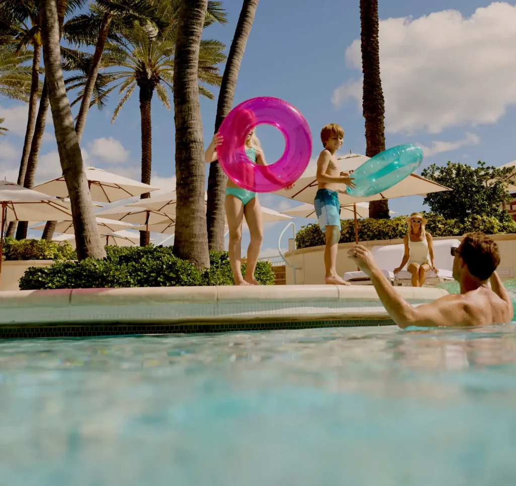 Family enjoying a sunny pool day with children holding colorful float rings among palm trees and white umbrellas