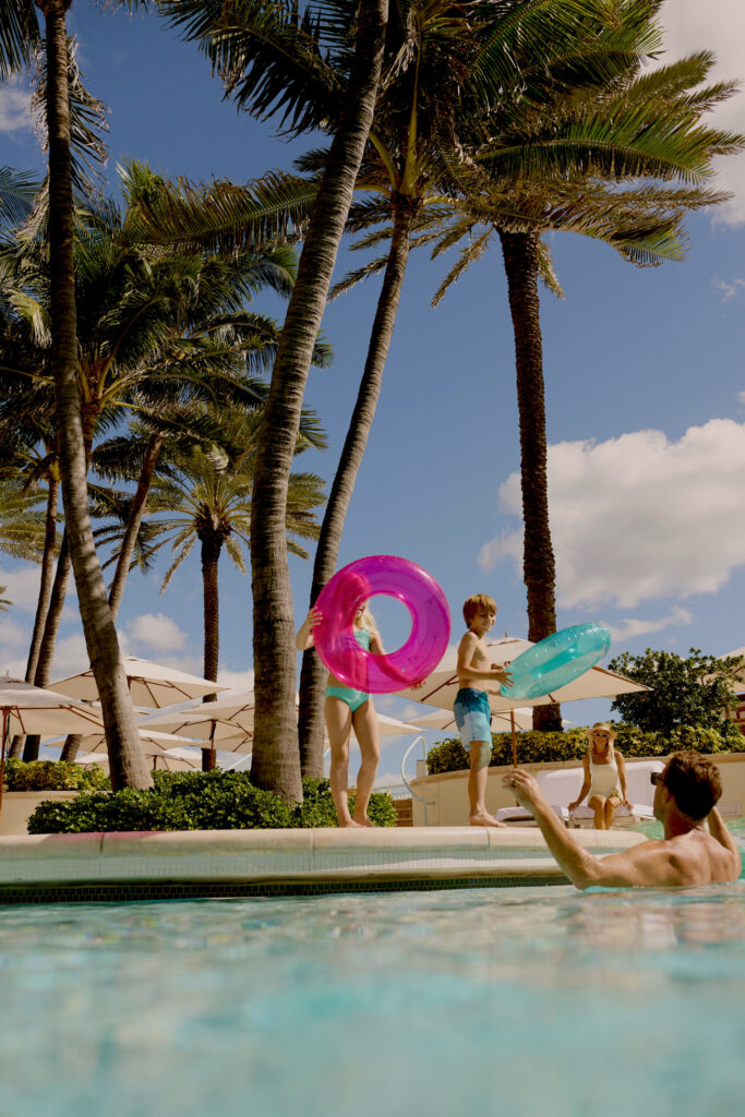 Children holding colorful pool floats by a resort pool with palm trees while a man reaches up from the water
