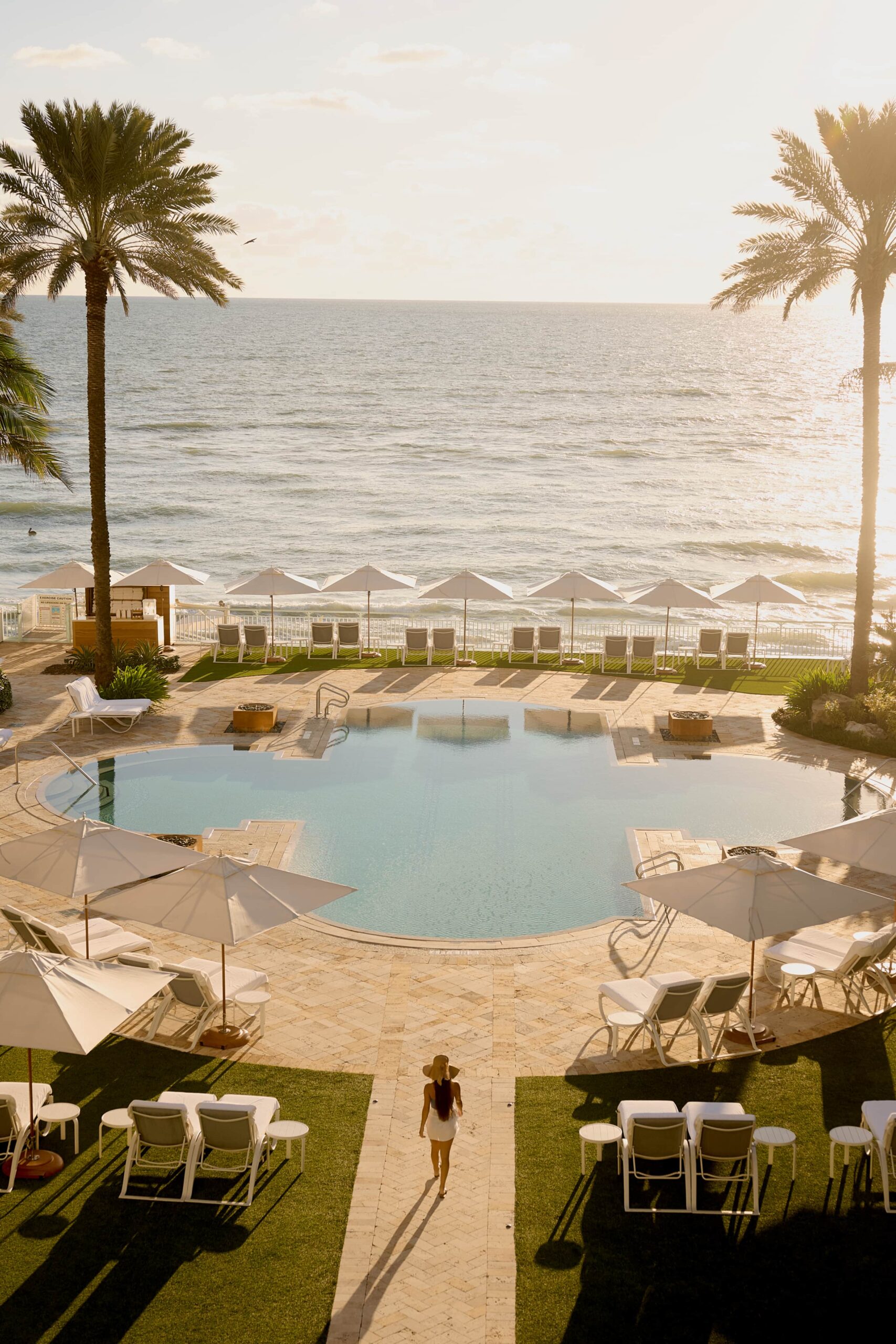 Oceanfront resort pool with lounge chairs, umbrellas, palm trees, and sunset view