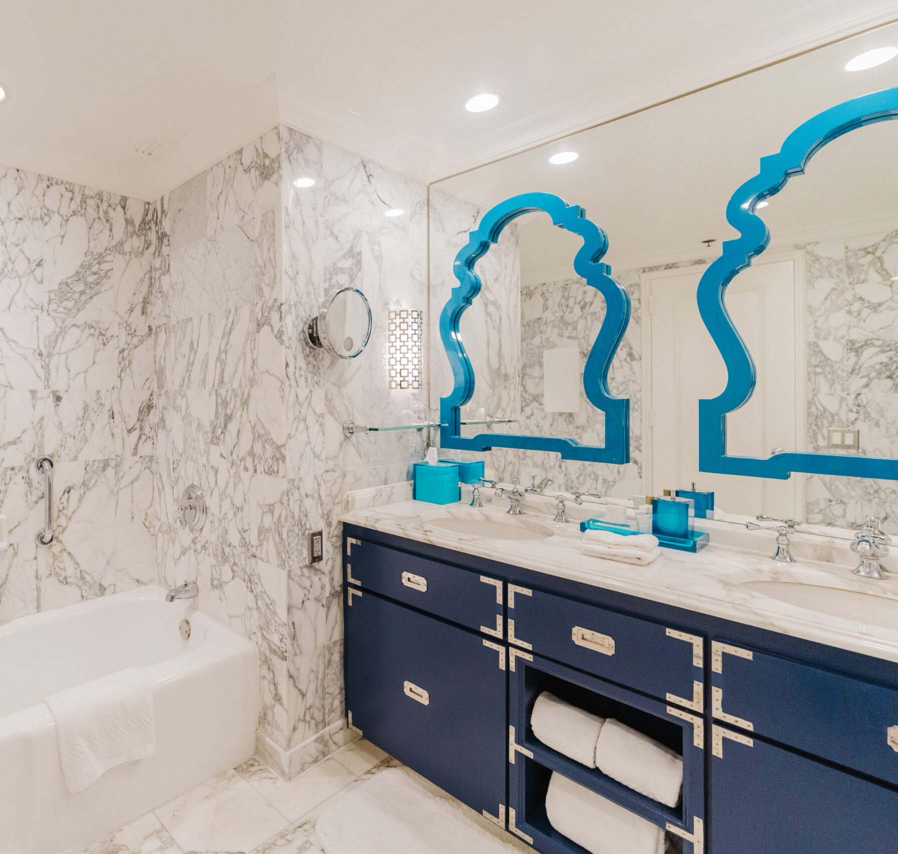 Modern marble bathroom with bathtub, blue vanity, dual sinks, and decorative mirrors under recessed lighting