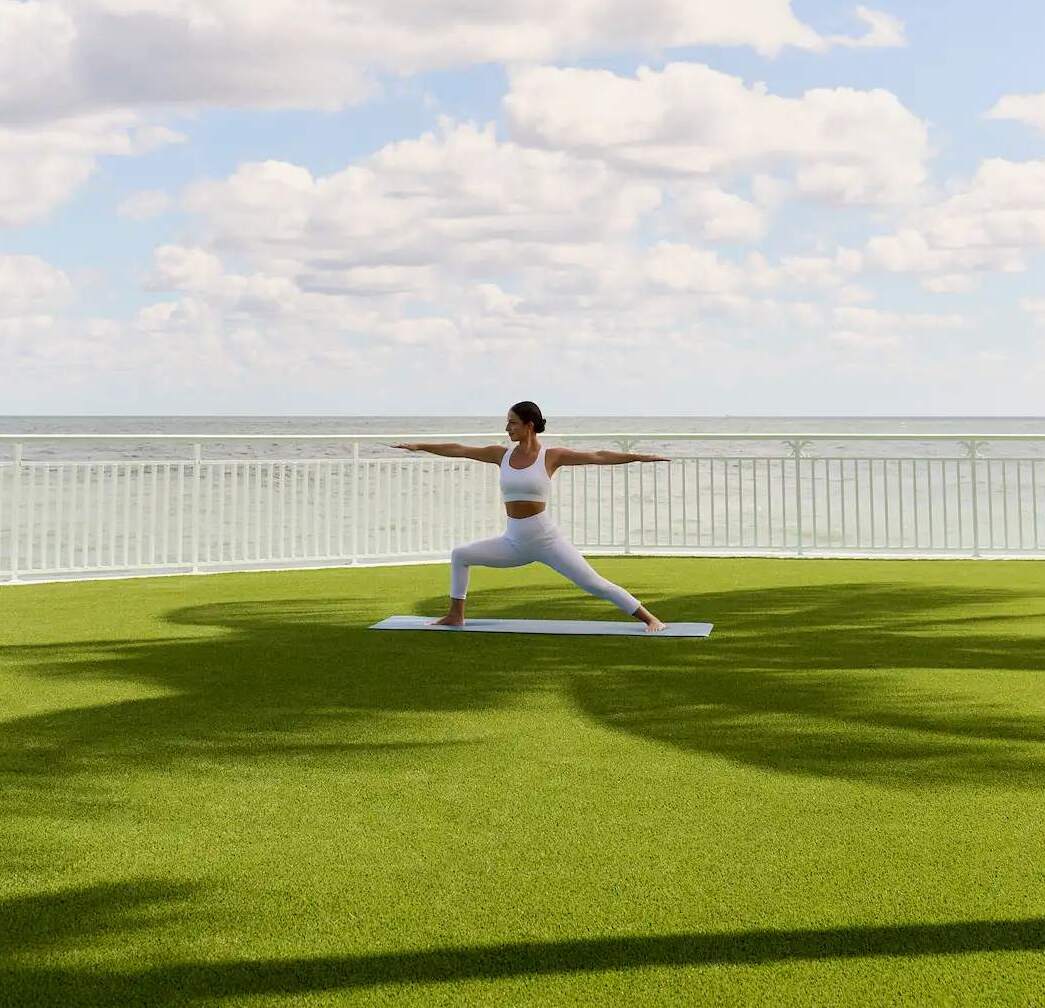 Person practicing Warrior II on a lawn overlooking the ocean under a bright sky and palm tree shade