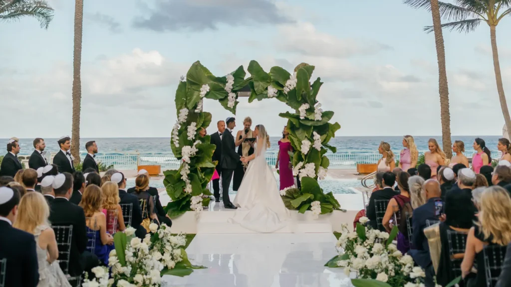 Bride and groom exchanging vows under floral arch at oceanfront wedding ceremony with guests seated nearby